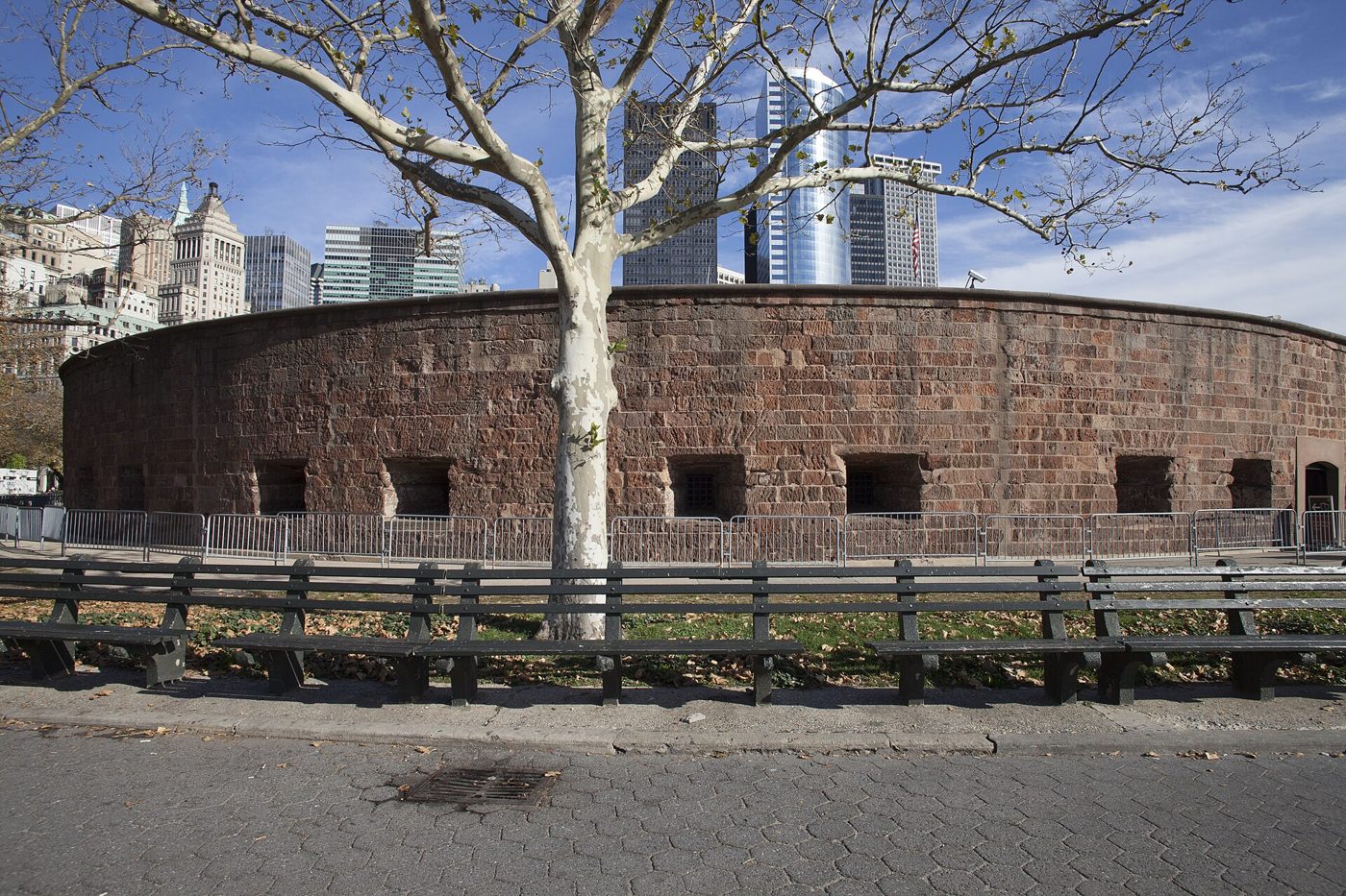 The round brownstone walls of Castle Clinton with square gun ports, seen with bare trees and downtown buildings behind