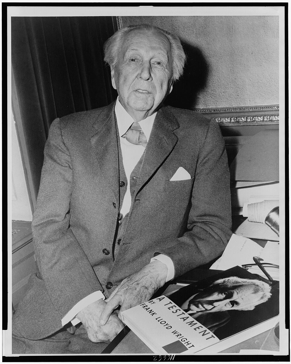 Frank Lloyd Wright seated in a suit, holding a newspaper, in a World-Telegram photograph