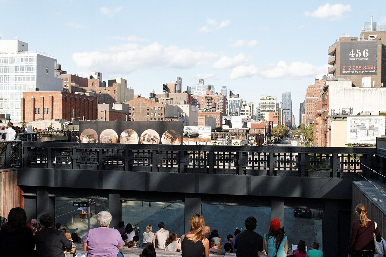 A crowd of visitors on the High Line along a railing and viewing area above a city street, with buildings and a tall tower in the distance