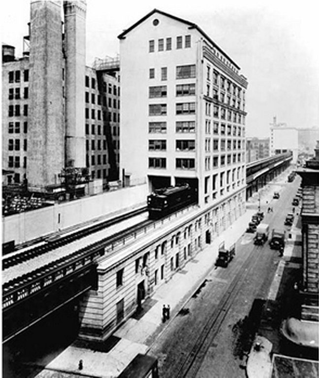 Black and white photograph of a tall industrial building with an elevated rail line and a train passing through at the level of the upper floors beside a street
