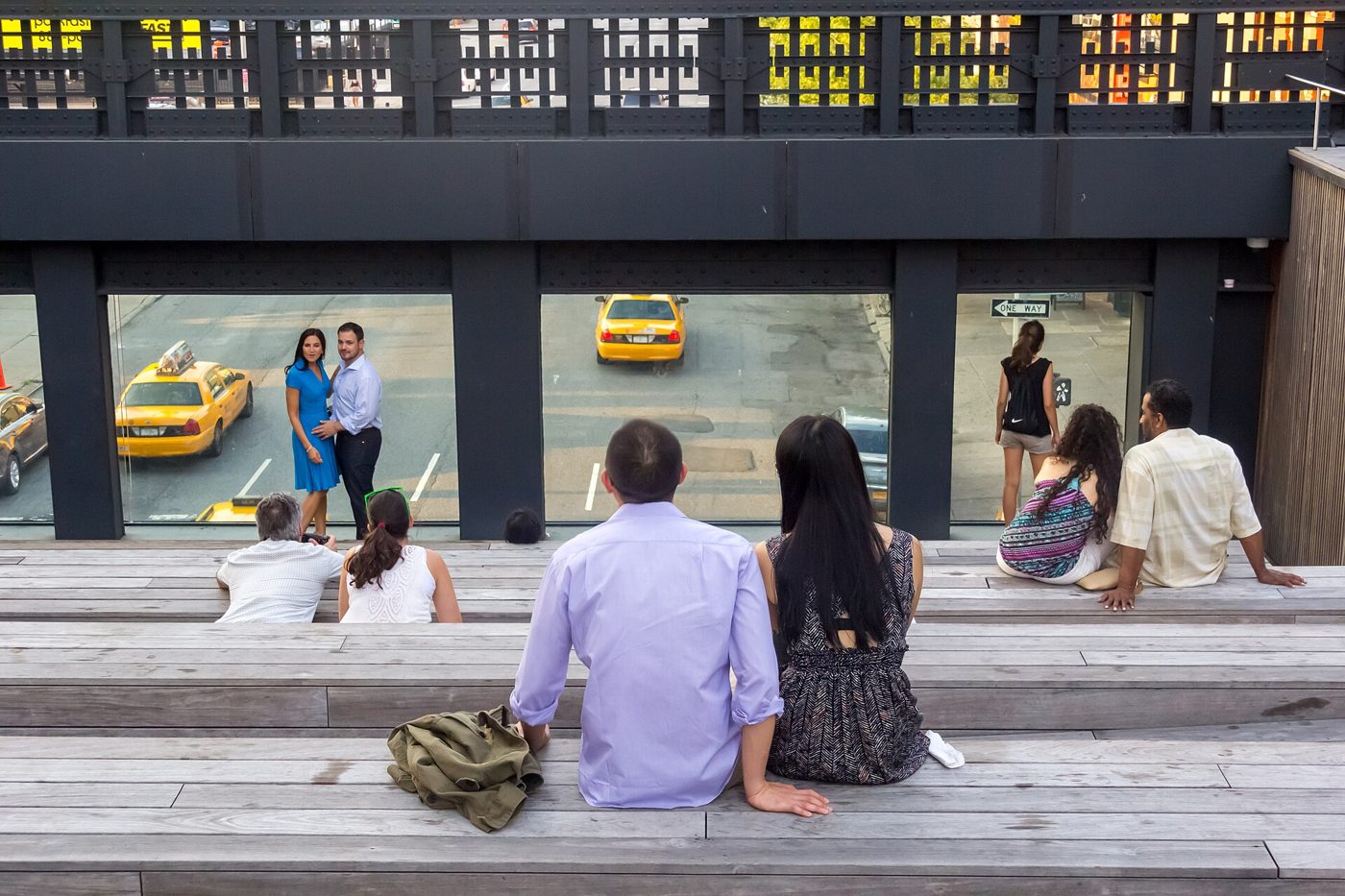 Visitors sitting on wide stepped wooden seating on the High Line looking down at a Chelsea street with yellow taxis