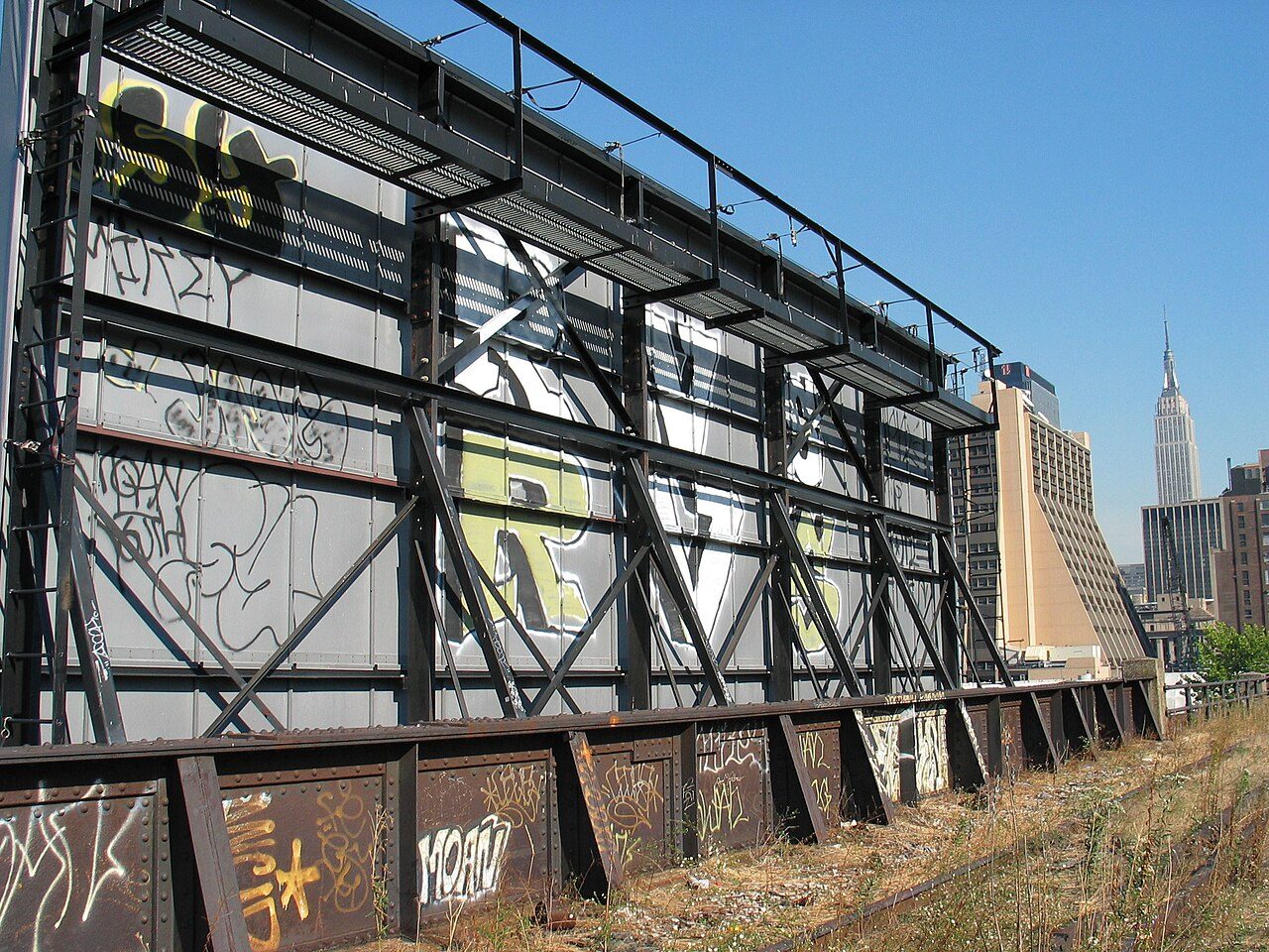 A weathered metal billboard frame beside a rail bed overgrown with grass on the abandoned High Line, with buildings in the distance