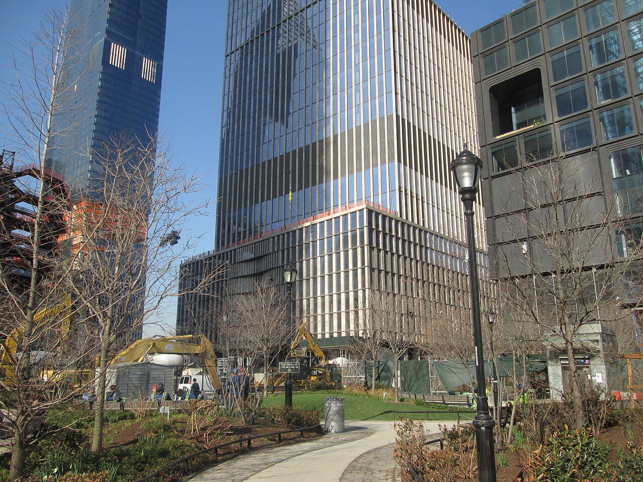 A plaza with young trees and a path in front of new glass and stone towers under construction on Manhattan West Side