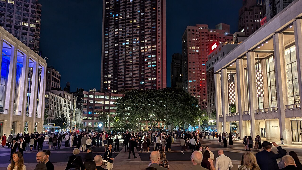 A crowd gathered on the Lincoln Center plaza at night, with illuminated halls on both sides and apartment towers behind