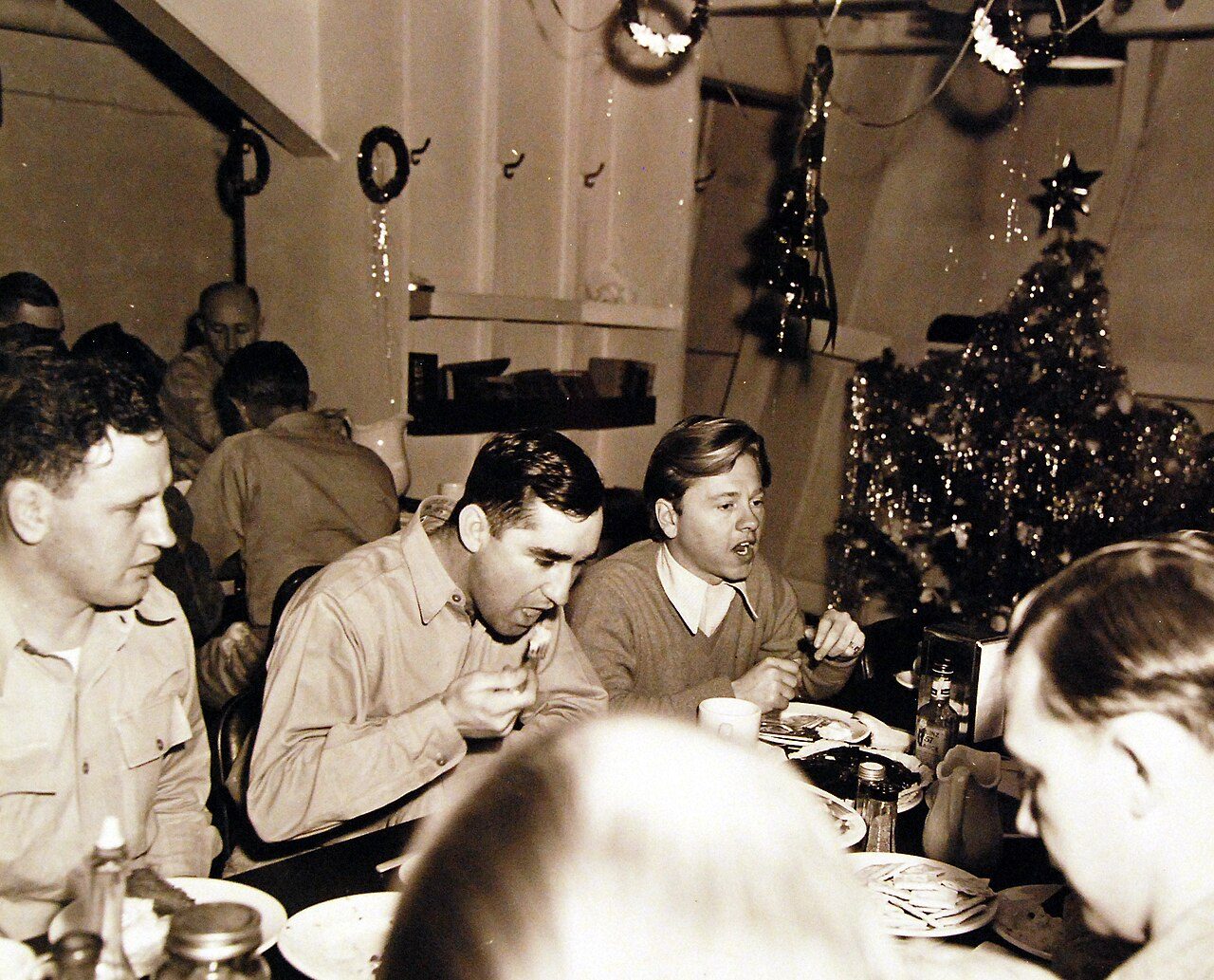 Sailors and a civilian entertainer seated at a crowded mess table beside a small decorated tree in the chief petty officers quarters of a warship