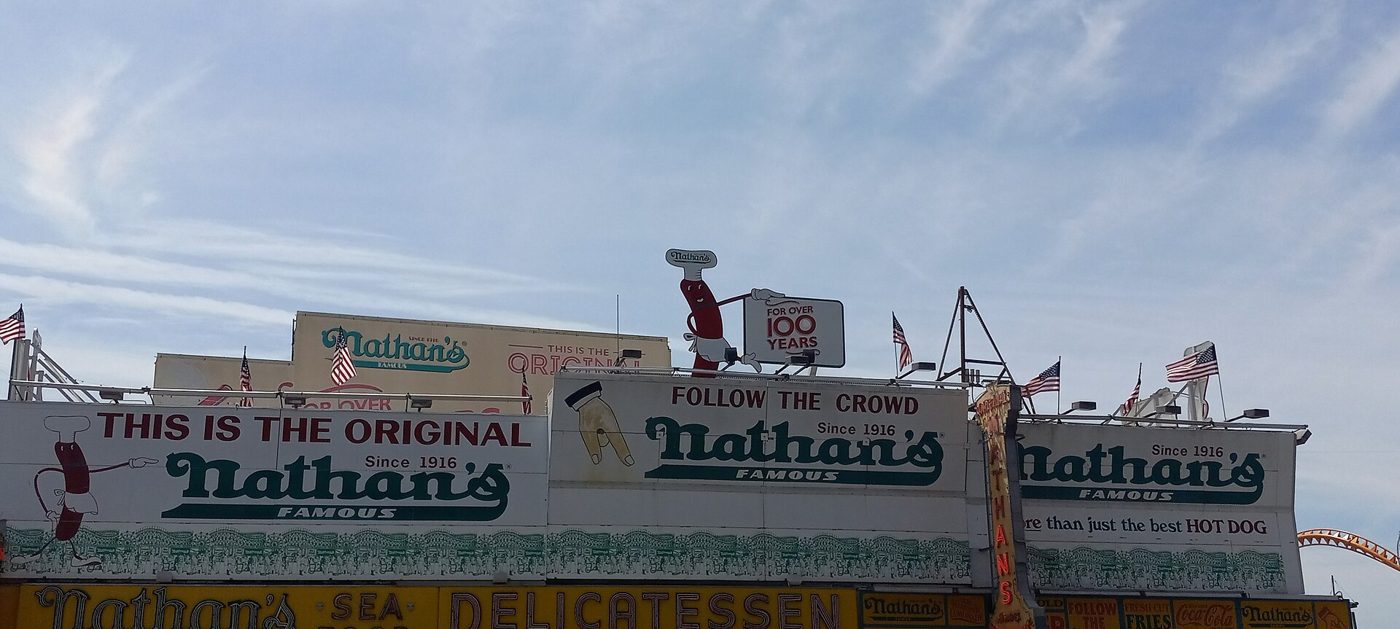 The white and red signs of Nathan's Famous on a long low building on Surf Avenue, with a blue sky behind