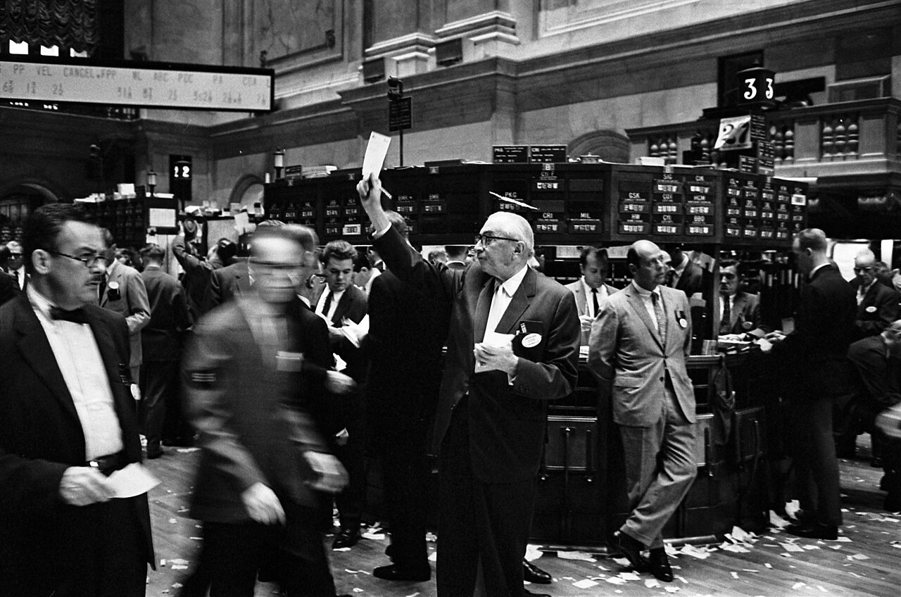 The NYSE trading floor around 1908, brokers in suits gathered at horseshoe-shaped trading posts under hanging annunciator boards