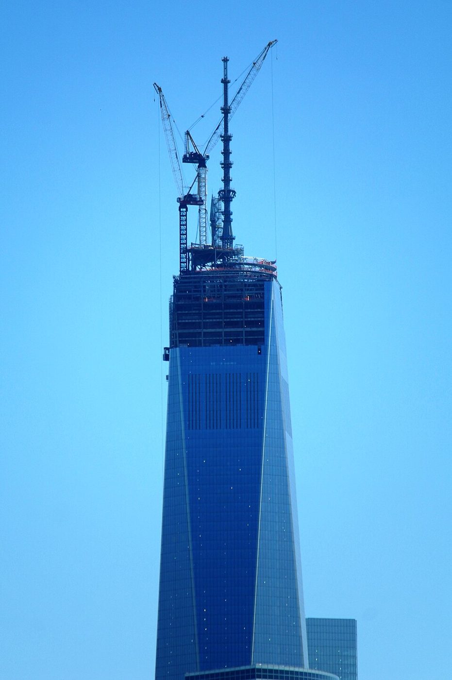 The top of the unfinished One World Trade Center against a blue sky, with cranes on the roof and the spire base in place
