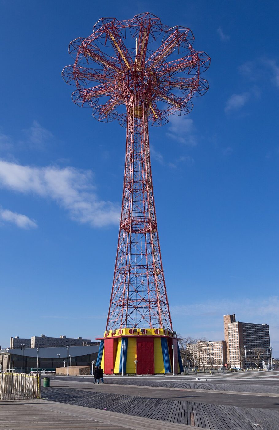 The red steel Parachute Jump tower on the Coney Island boardwalk against a blue sky, with colorful low buildings at its base