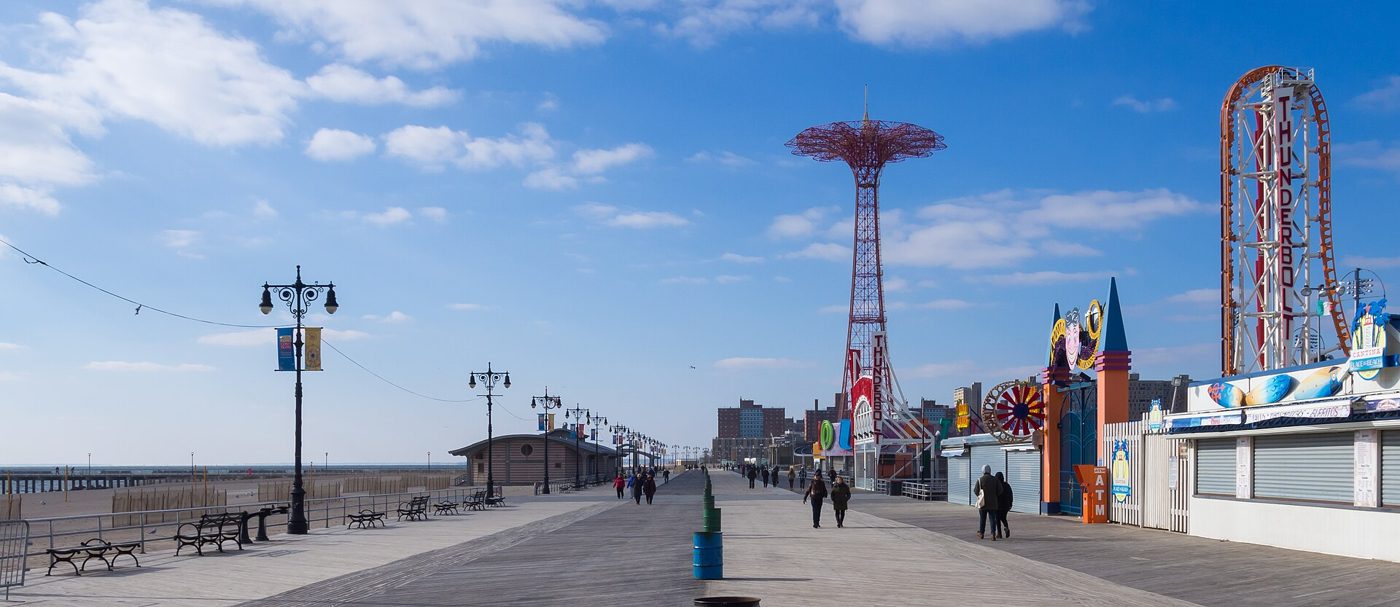 A wide wooden boardwalk on a sunny day with lamp posts, the red Parachute Jump tower and amusement rides in the distance