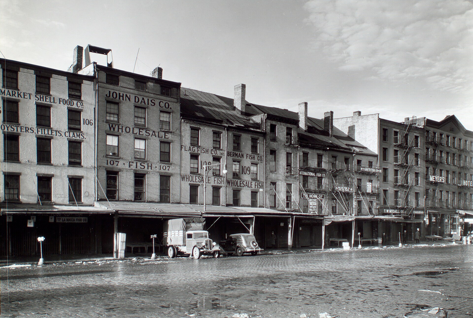 Berenice Abbott's 1935 photograph of fish dealers' storefronts and the Meyers Hotel in the low buildings along South Street, with a truck and a car on the cobblestones