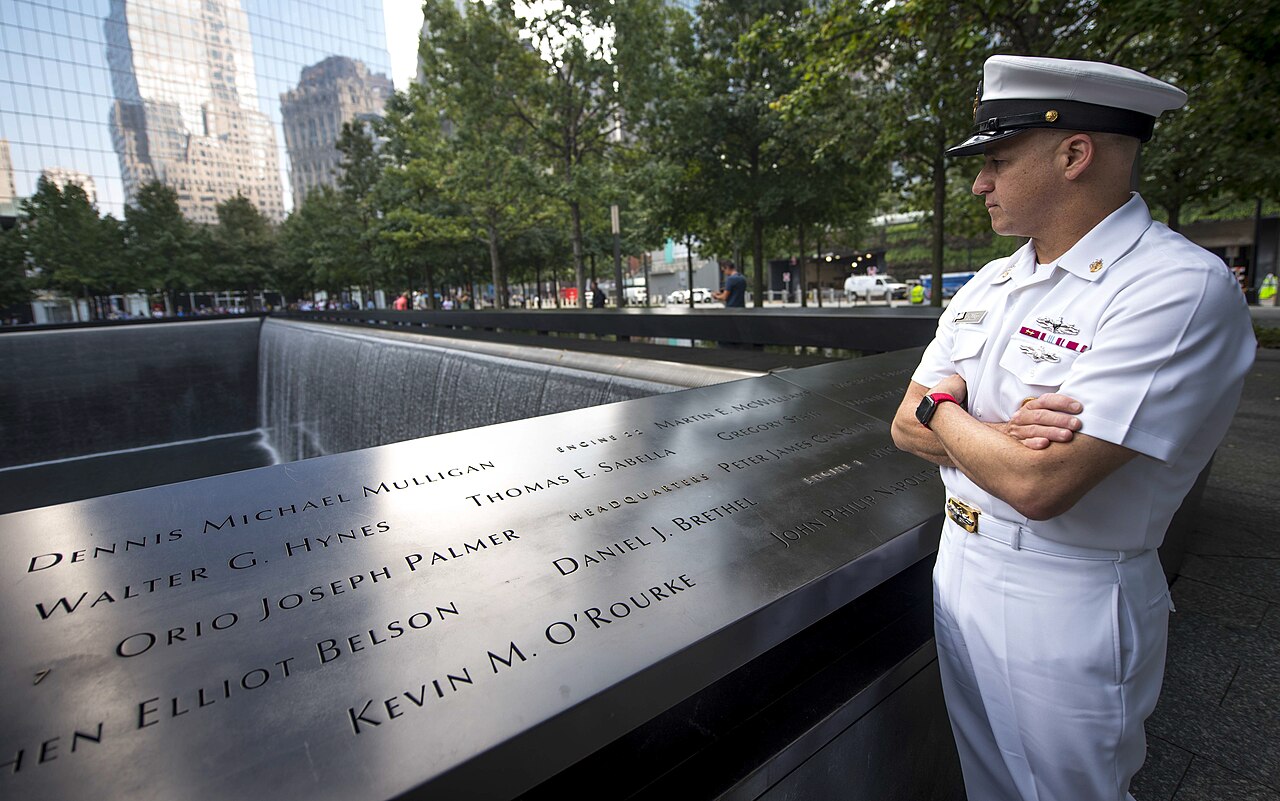 A US Navy sailor in dress whites standing at the bronze names parapet of the memorial, the pool and plaza trees behind him
