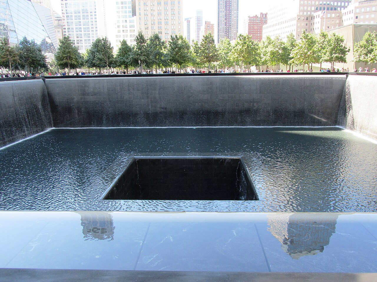 A 9/11 Memorial reflecting pool with water sheeting down its walls into a central square void, oak trees and Lower Manhattan towers beyond