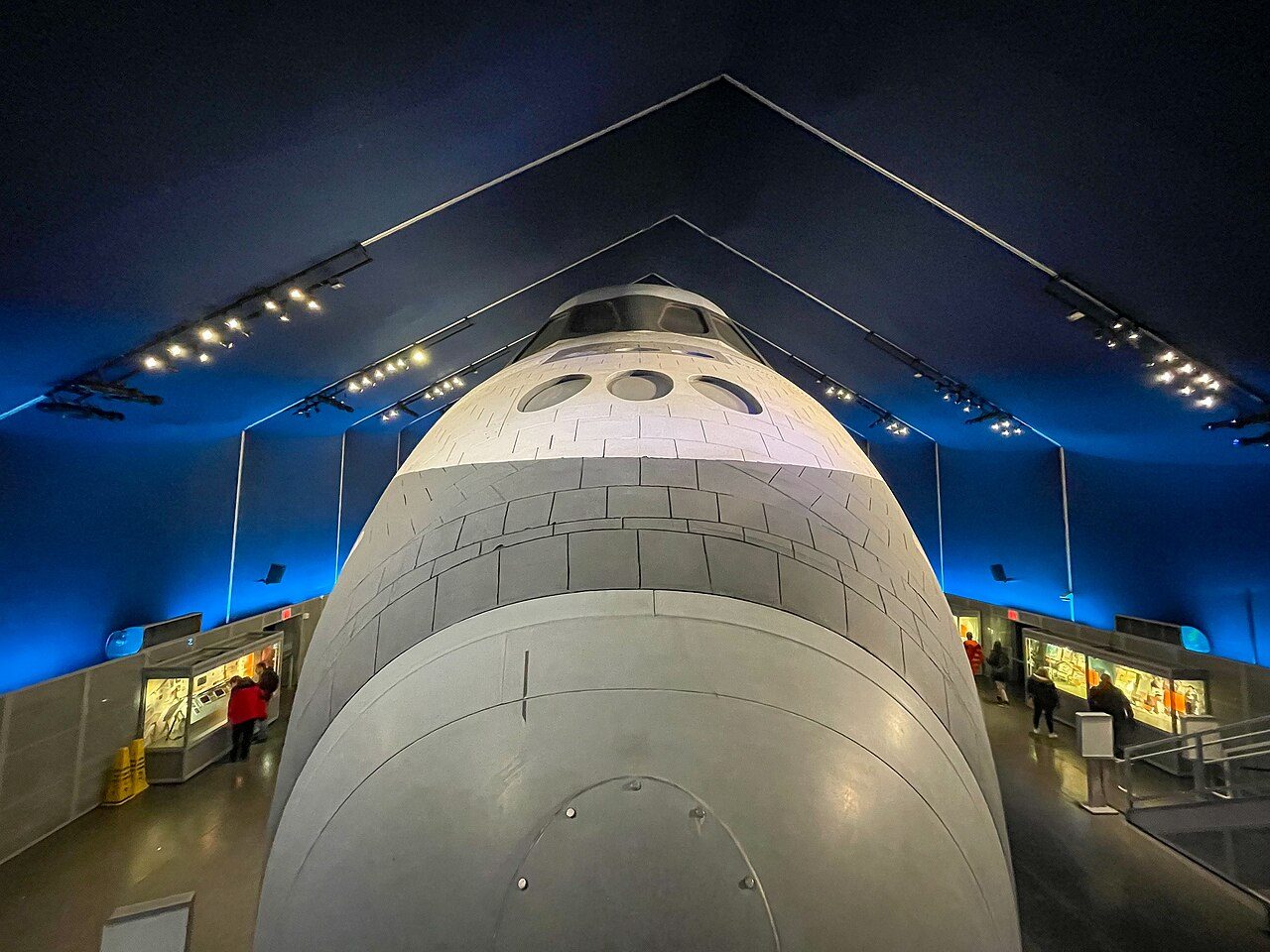 Looking down on the white nose and cockpit windows of the Space Shuttle Enterprise inside a dark blue pavilion