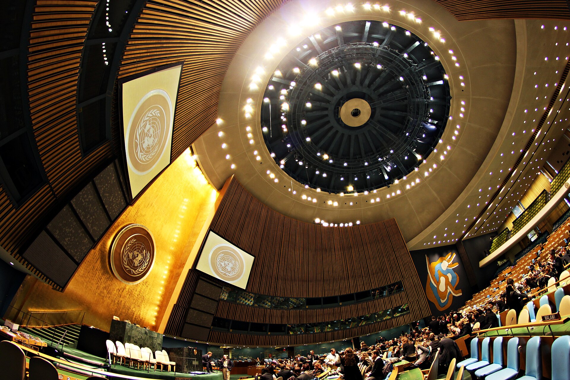 The interior of the UN General Assembly Hall, looking up into its illuminated dome, with the UN emblem and a colorful mural on the walls