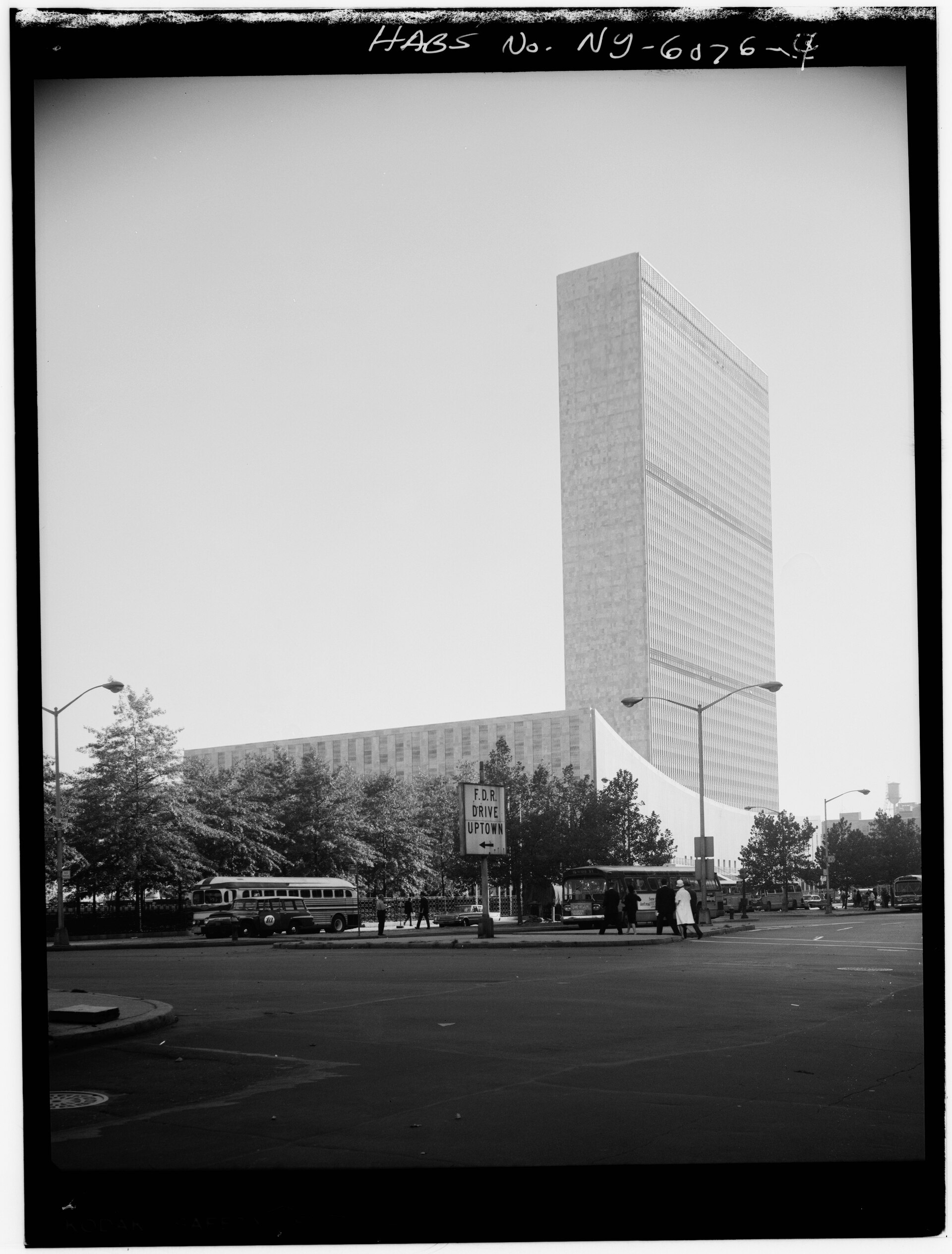 A mid-century black and white photograph of the marble Secretariat tower rising behind the low General Assembly building, with buses on the street below