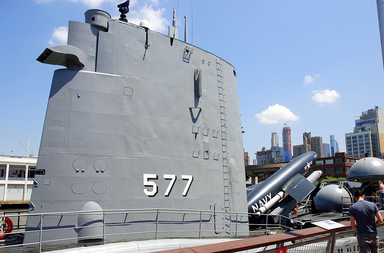 The grey sail of the submarine Growler with the number 577 painted on it, beside the pier with Midtown buildings behind