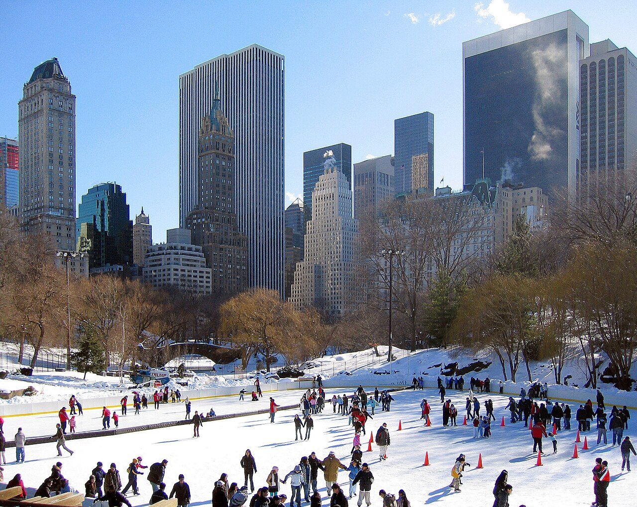 Skaters on the ice at Wollman Rink in winter, with Midtown Manhattan towers rising behind bare trees